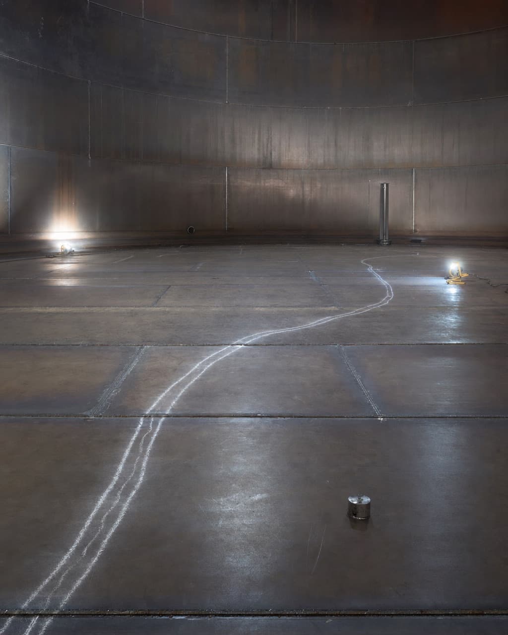 The cleaned bare steel floor plates inside an empty storage tank, lap welds visible, lit by portable work lamps