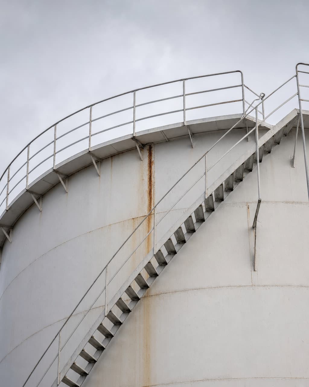 The external stairway and upper shell of a storage tank against a grey sky, with rust bleed at a seam